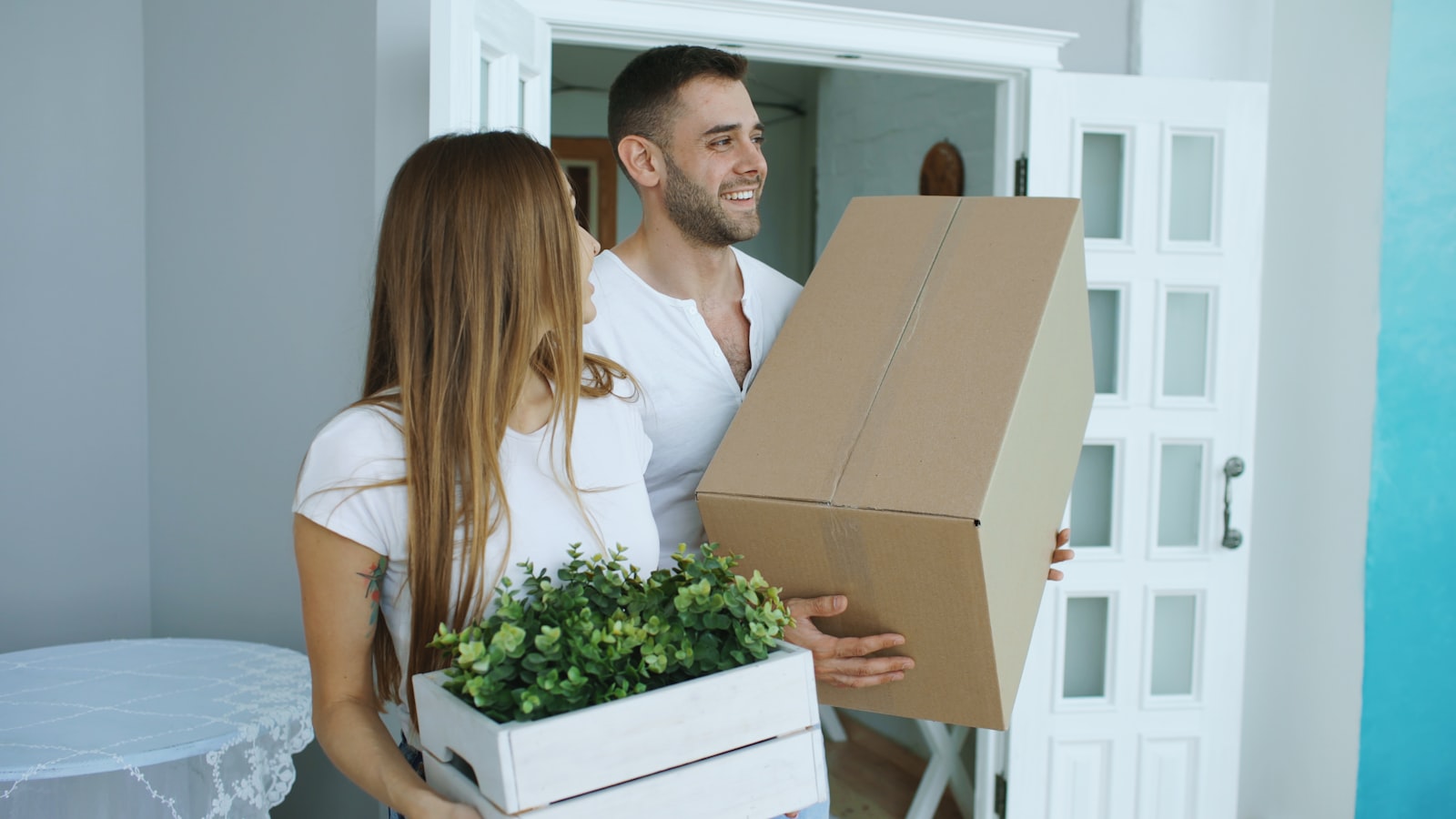 A couple carrying cardboard boxes and a plant through the door of a new home