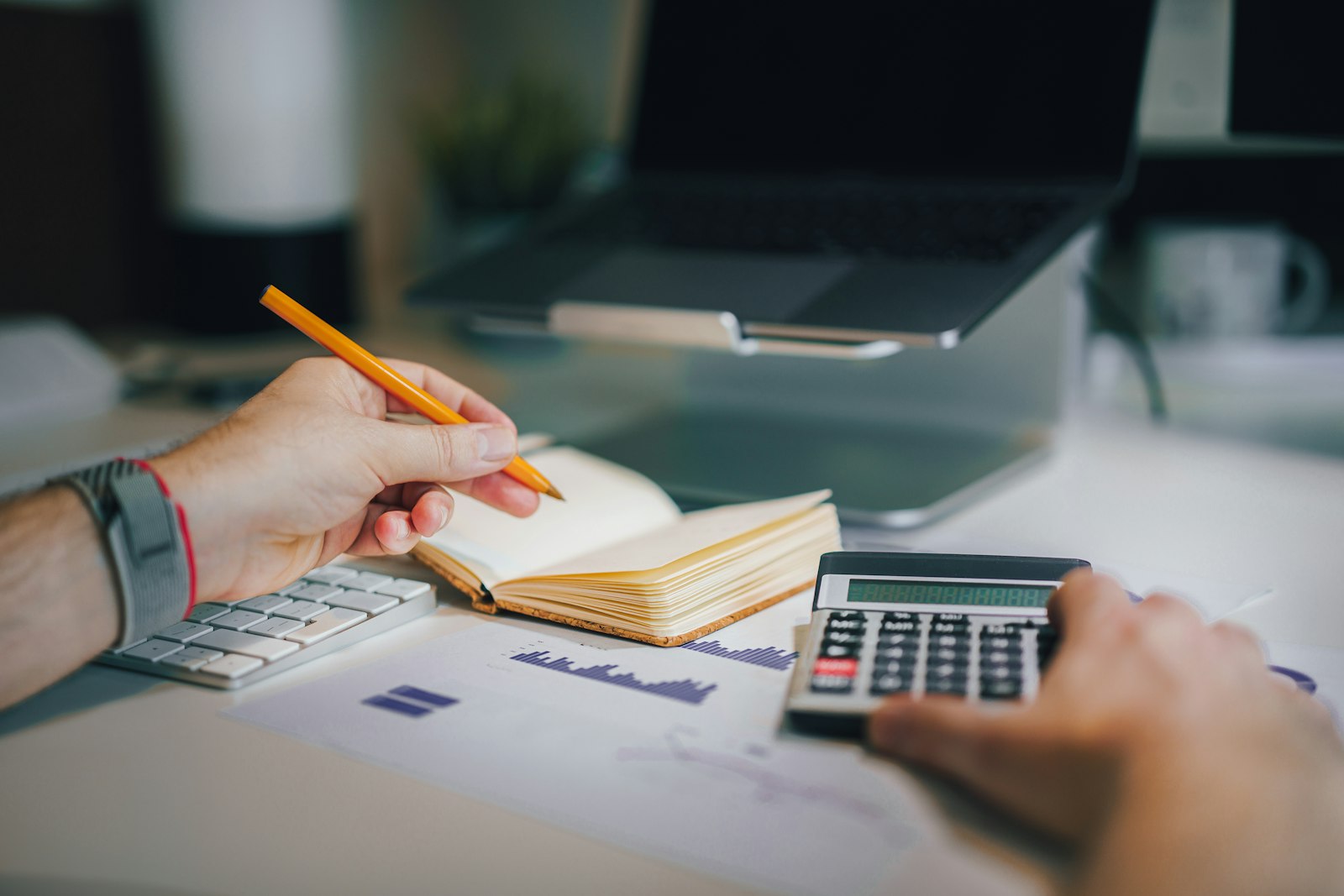 Hands using a calculator beside an open notebook, a printed chart and a laptop