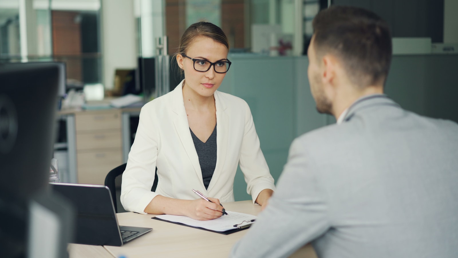 An interviewer taking notes across a desk from a candidate in an office