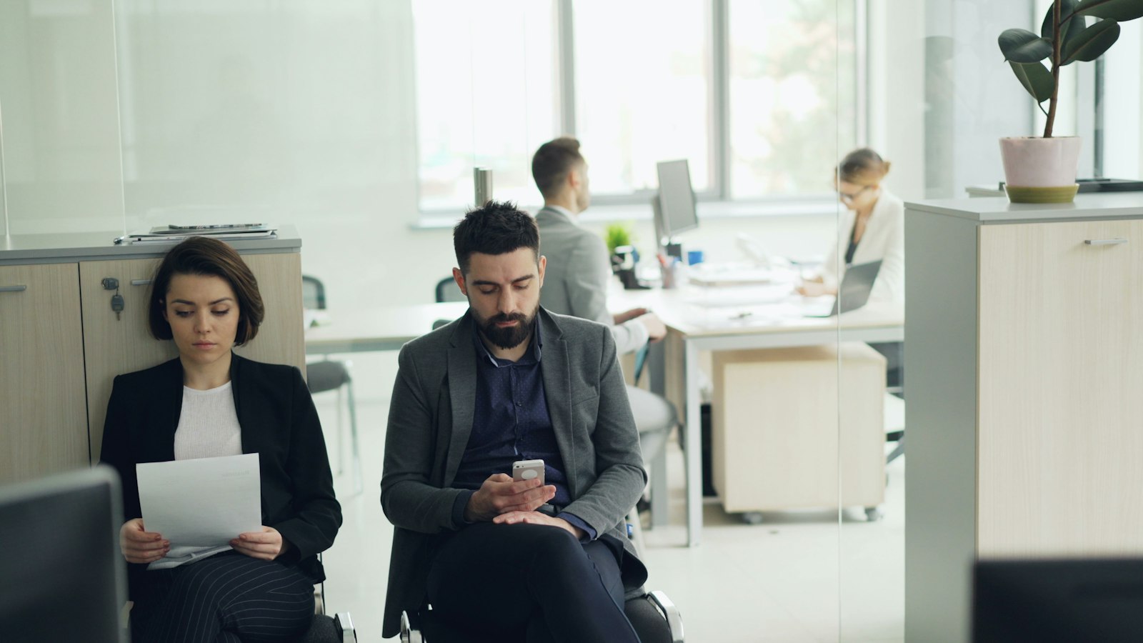 Two candidates waiting in an office, one holding a printed resume
