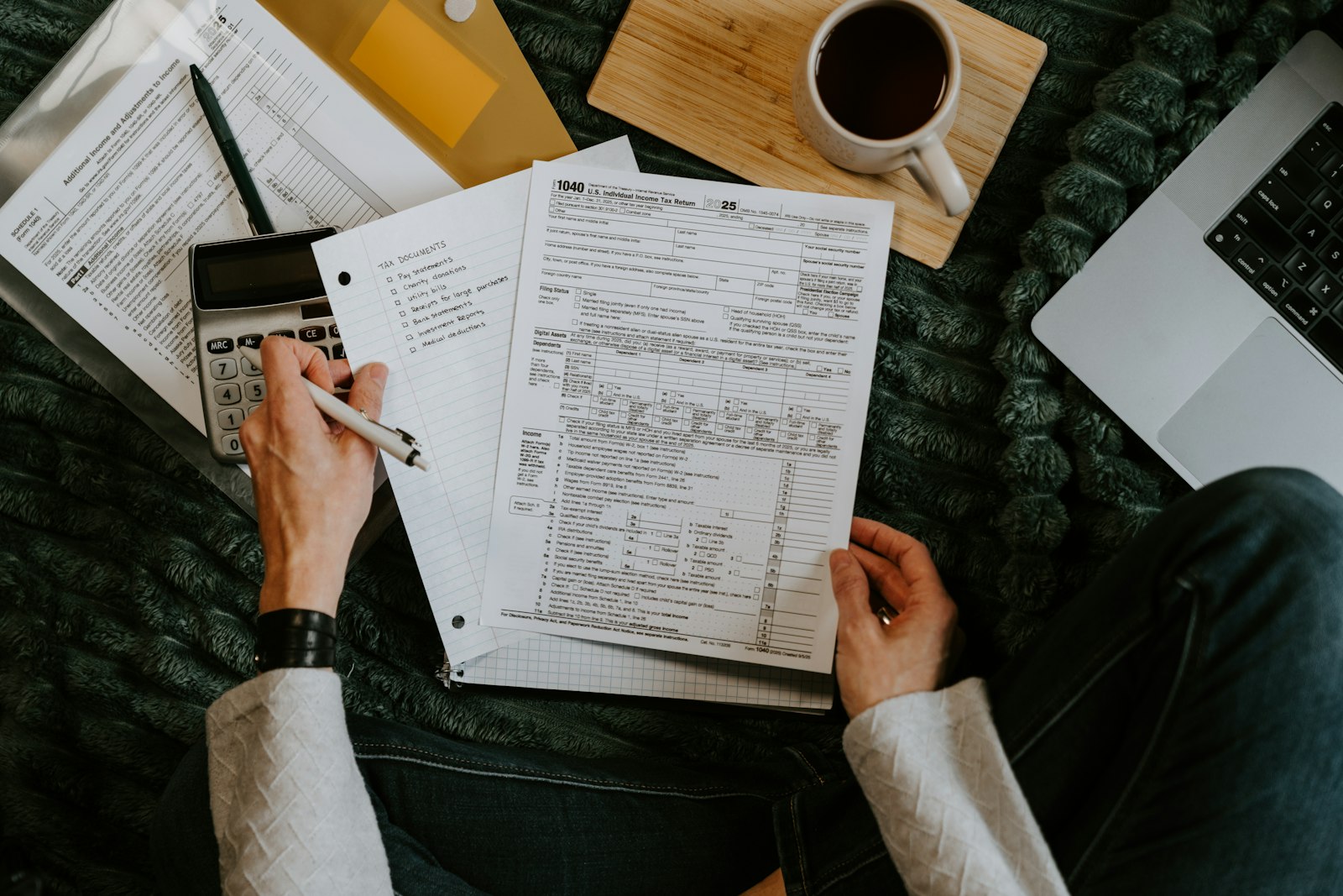 A person filling in a Form 1040 tax return beside a calculator and a handwritten tax documents checklist